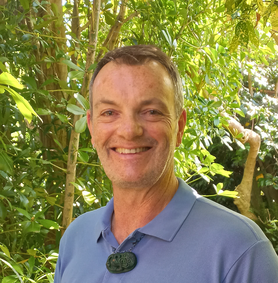 Richard Shaw wearing a blue shirt and a pounamu standing in front of green foliage